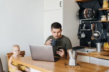 Man Father works from home at the laptop, sitting in the interior of the kitchen at the dining table with a small child. quarantine isolation Coronavirus.