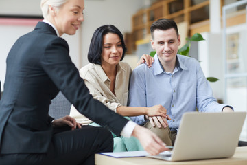 Mature real estate agent using laptop and showing to young couple some presentation of their new house