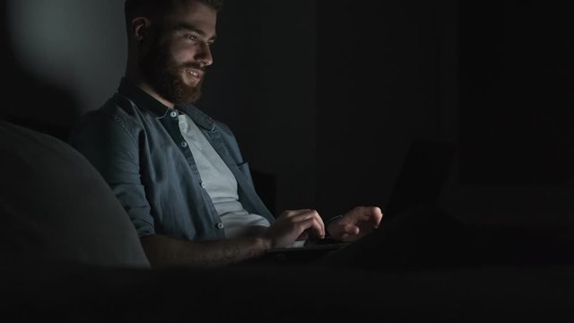 A side view of a handsome young man is using his laptop computer at night time in the dark room