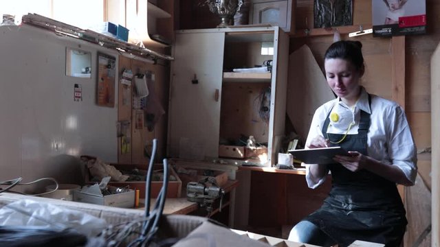 Confident woman working as carpenter in her own woodshop. She using a tablet pc and writes notes while being  in her workspace. Small business concept.