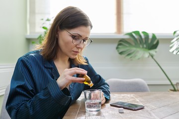 Mature woman dripping drops with glass of water, drinking sedative medicine