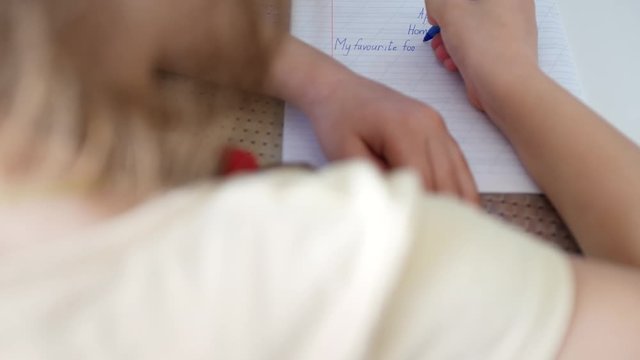 Close-up hand of a child schoolgirl who does homework at an English lesson. In a notebook with slanting lines, she carefully writes English letters and words with a pen