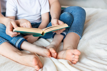 A child and a young woman are sitting on the bed and reading a fairy tale. An open book lies on its knees. Home leisure - reading a book.