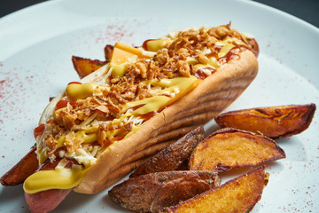 Appetizing classic American hot dog with caramelized onions, cheddar cheese, mustard and ketchup with a side dish of potatoes on a white background. Fast food