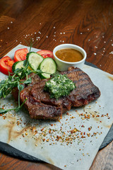 Juicy and mouth-watering grilled ribeye steak with spicy butter on a black tray with a side dish of vegetables. American cuisine.. Close up. Wooden background