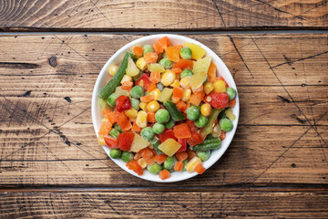 Chopped frozen vegetables in a plate on a wooden background. Corn peas pepper carrots. copy space.