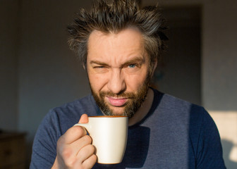 A funny man with a beard after waking up with a large cup of coffee or tea looks at the camera with one eye.