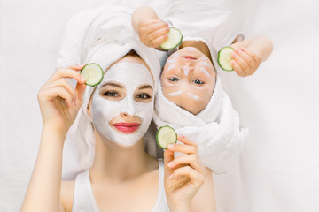 Happy family spa procedures, mothers day. Pretty young mother and baby girl toddler lying head to head on isolated white background, with face skin mask and towels on head, holding slices of cucumber