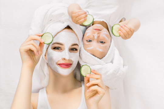 Happy family spa procedures, mothers day. Pretty young mother and baby girl toddler lying head to head on isolated white background, with face skin mask and towels on head, holding slices of cucumber