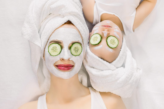 Mother and little daughter having spa procedures together. They are in white bath towels on head and with slices of cucumber on their eyes. Woman has white facial mask on her skin