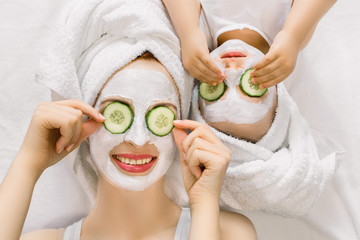 Photo of cheerful mother and little daughter with wrapped hair in white bath towels, white facial mud mask on faces, lying on the bed, and applying pieces of cucumber to their eyes. Family spa