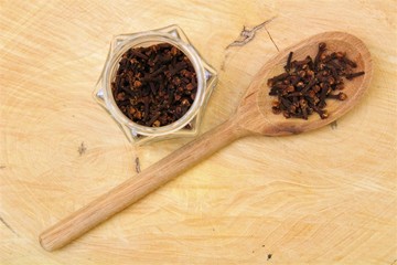 Top view of cloves (Syzygium aromaticum) on a wooden spoon and inside a glass pot on raw wooden board.