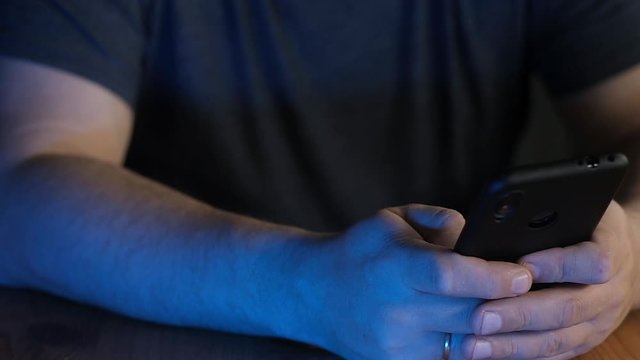 adult man is sitting in an office at a workplace and writing a message on a smartphone. Close-up of a man's hand dialing a number on a cell phone in neon light. Modern wireless technology.