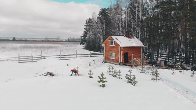 Aerial view. A Caucasian man holding an axe, chopping wood in winter. A man stands in the courtyard of a country house, which is located next to the forest. The camera rises from the bottom up.