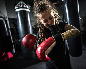 Girl puts on boxing gloves in gym. Girl with tattoos and dreadlocks wears boxing gloves.
