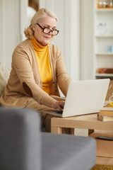 Senior woman in eyeglasses sitting on sofa and working online she typing on laptop computer at home