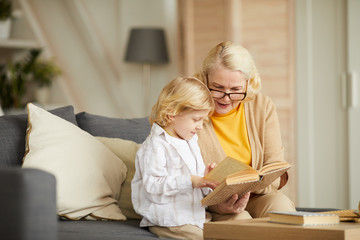 Grandmother wearing eyeglasses and reading a book to her grandson while they sitting on sofa in the...