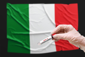 A doctor holds a test tube with a positive blood test for coronavirus in front of the Italian flag. Covid-19 epidemic, infection, pandemic, pneumonia.