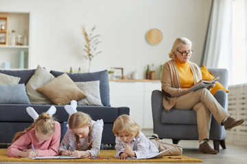 Children lying on the floor and drawing with pencils with their grandmother sitting on armchair and...