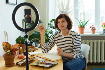 Female artist filming design workshop at home for online education. Working from home making master-class using ring lighting. Cozy artist's studio.