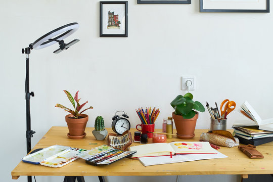 Artist's workplace for working from home with watercolor paints, brushes and sketchbooks. Place for design, illustration and creativity. Blogger ring light standing next to the table.