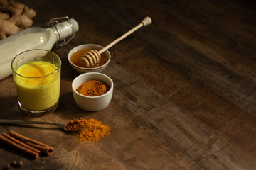 Turmeric Milk is poured into a glass. Next to it are the ingredients for making a drink - milk in a bottle on a napkin, spices and honey. Golden drink on dark moody background, selective focus. Top