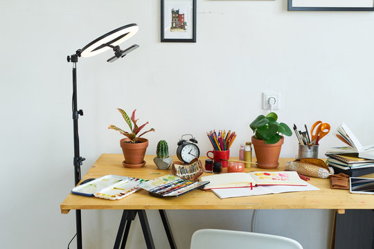 Artist's workplace for working from home with watercolor paints, brushes and sketchbooks. Place for design, illustration and creativity. Blogger ring light standing next to the table.