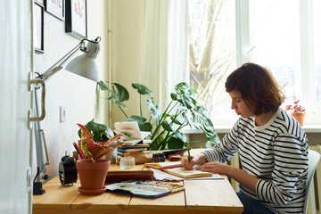 Female artist at her workplace working from home. Woman dressed in jeans and striped shirt sitting...