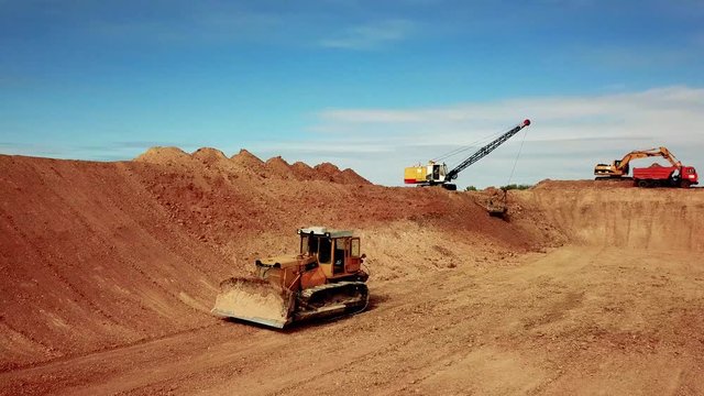 Drone wide shot from right to left inside the construction site following the bulldozer moving forward. Behind, one excavator digging the sand, another fill a dump truck model Kamaz with sand.