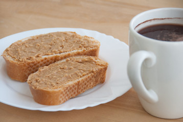 White plate with peanut butter toasts and a cup of coffee on the wooden table. Close-up