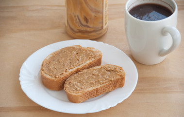 Toasted bread and peanut butter on a white plate, a cup of coffee and jar on the wooden table close-up
