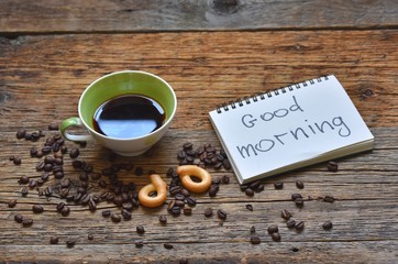 Fresh Cup of brewed coffee on a wooden background and a greeting note in the notebook Good Morning 
