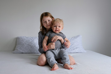 girl and boy play on a white bed at home in isolation. brother and sister have fun