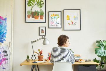 Female artist at her workplace working from home. Young woman dressed in jeans and striped shirt sitting at the table turned backwards. Creating an illustration.