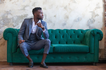 Stylish African black man is sitting on a green sofa in a studio in three piece suit against a gray concrete wall