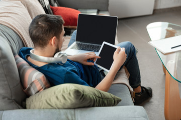 Young focused man studying at home during online courses or free information by hisself. Becomes software developer, IT manager while isolated, quarantined. Using laptop, smartphone, headphones.