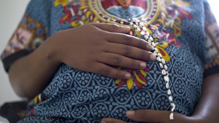 Close up. Third trimester pregnant African woman, in traditional clothing, cradles her belly at rural hospital.
