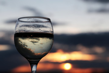 Glass with white wine on beautiful sunset background, sun and sky are reflected in alcohol drink. Concept of celebration, evening party at resort, romantic dinner outdoors