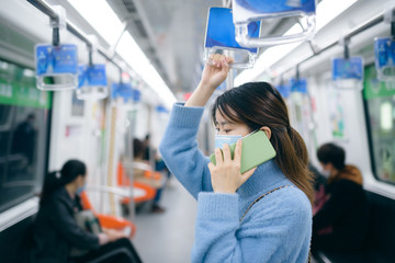 That young Asian woman wearing a surgical mask is using smart phone in the subway train.