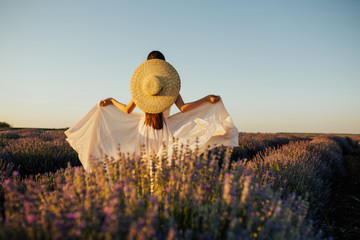 Obraz premium Free brunette woman with long hair and with open arms enjoying sunny day in lavender field. Harmony. Attractive girl with straw hat and tender dress dreaming. Blossoming lavender field.