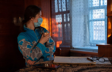 Fototapeta na wymiar girl sits at home on a quarantine face mask and drinks from a cup. Looks out the window on the scribble table