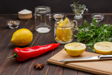 Sliced lemon and knife on cutting board. Lemon in jar. Chili pepper, whole lemon and salt on table.