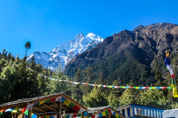 Colorful prayer flags waving above the village's rooftops. In the back high Himalayan peaks seen from Annapurna Circuit Trek, Nepal. Meditation and peace of mind. High snow caped mountains peaks.