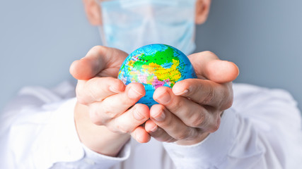 Close-up of a man's hands holding a small globe, a man in a medical mask out of focus, dressed in a white shirt, uniform on a gray background.   Coronavirus, Covid-19