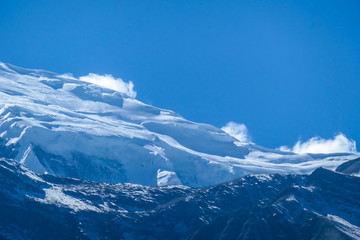 A close up view on snow caped Himalayan peak seen from Annapurna Circuit Trek, Nepal. Sharp and steep slopes of the mountain. Powder snow being blown by strong wind. First sunbeams reaching the peak