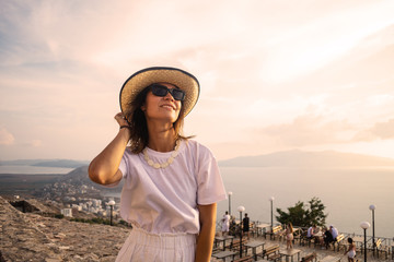 Fototapeta premium Young woman in hat, white t-shirt and shorts standing on the stone, looking at sunset over the sea.