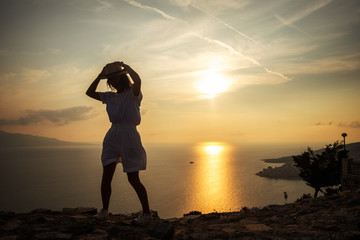 Fototapeta premium Young woman in hat, white t-shirt and shorts standing on the stone, looking at sunset over the sea.