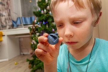A sad little European boy looks at a spoiled bad blue ball for the Christmas tree. Preparing for the New year, decorating the Christmas tree.