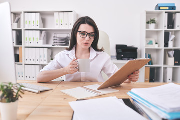 Pretty young female accountant in eyeglasses having tea and reading paper