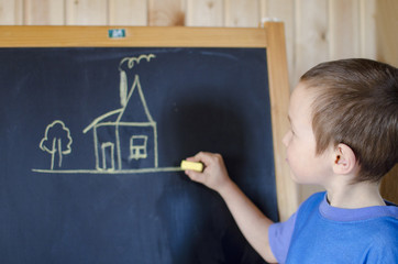 a boy in a blue t shirt at the blackboard drew a house with a chimney and smoke with chalk